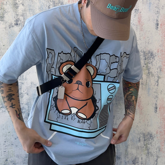 Person wearing a light gray t-shirt with a cartoon bear design and a brown cap, standing against a textured wall.